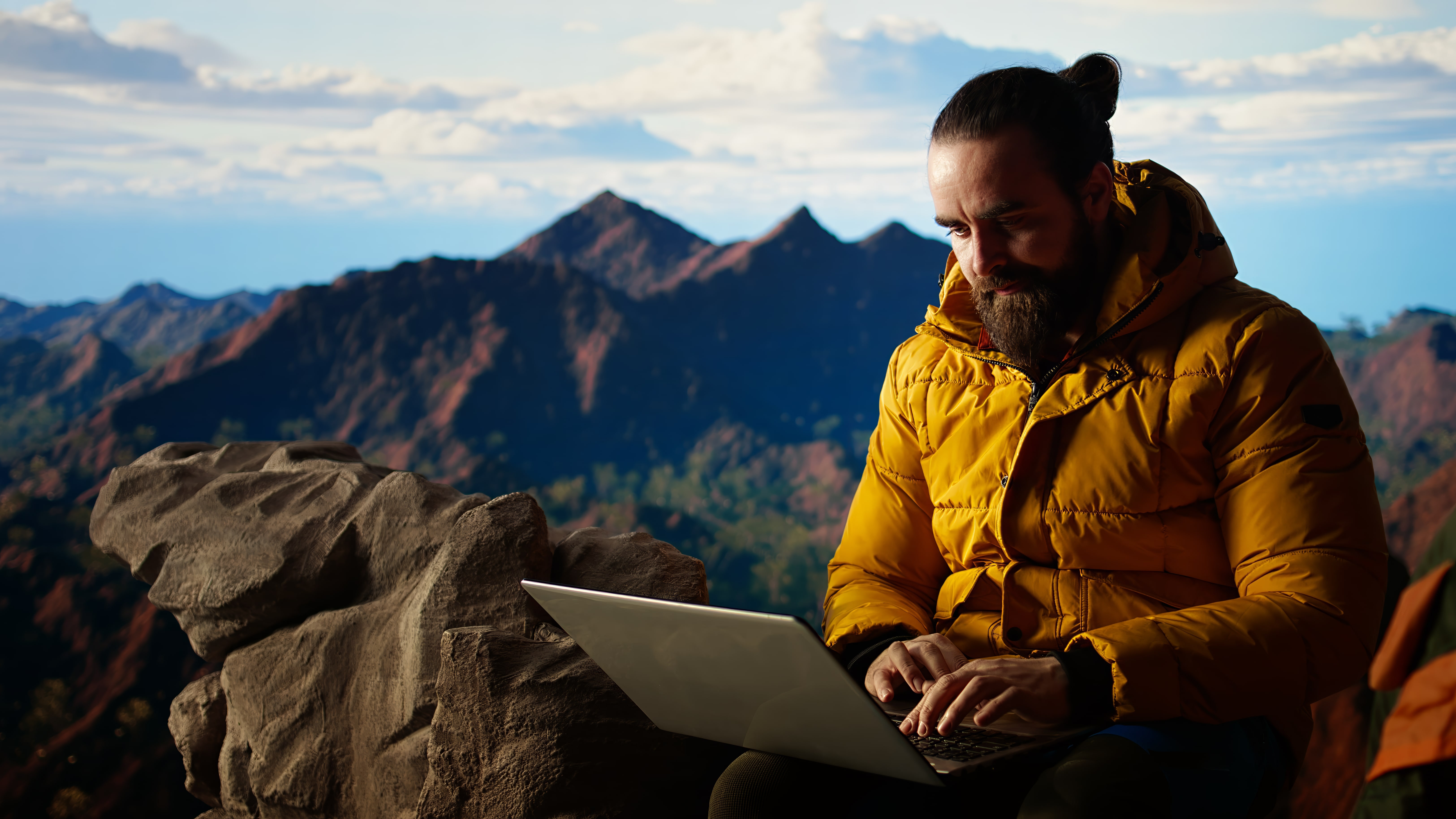 Outdoor-Arbeiten mit Laptop in den Bergen: Beleuchteter digitaler Nomade in gelber Jacke genießt die Natur.