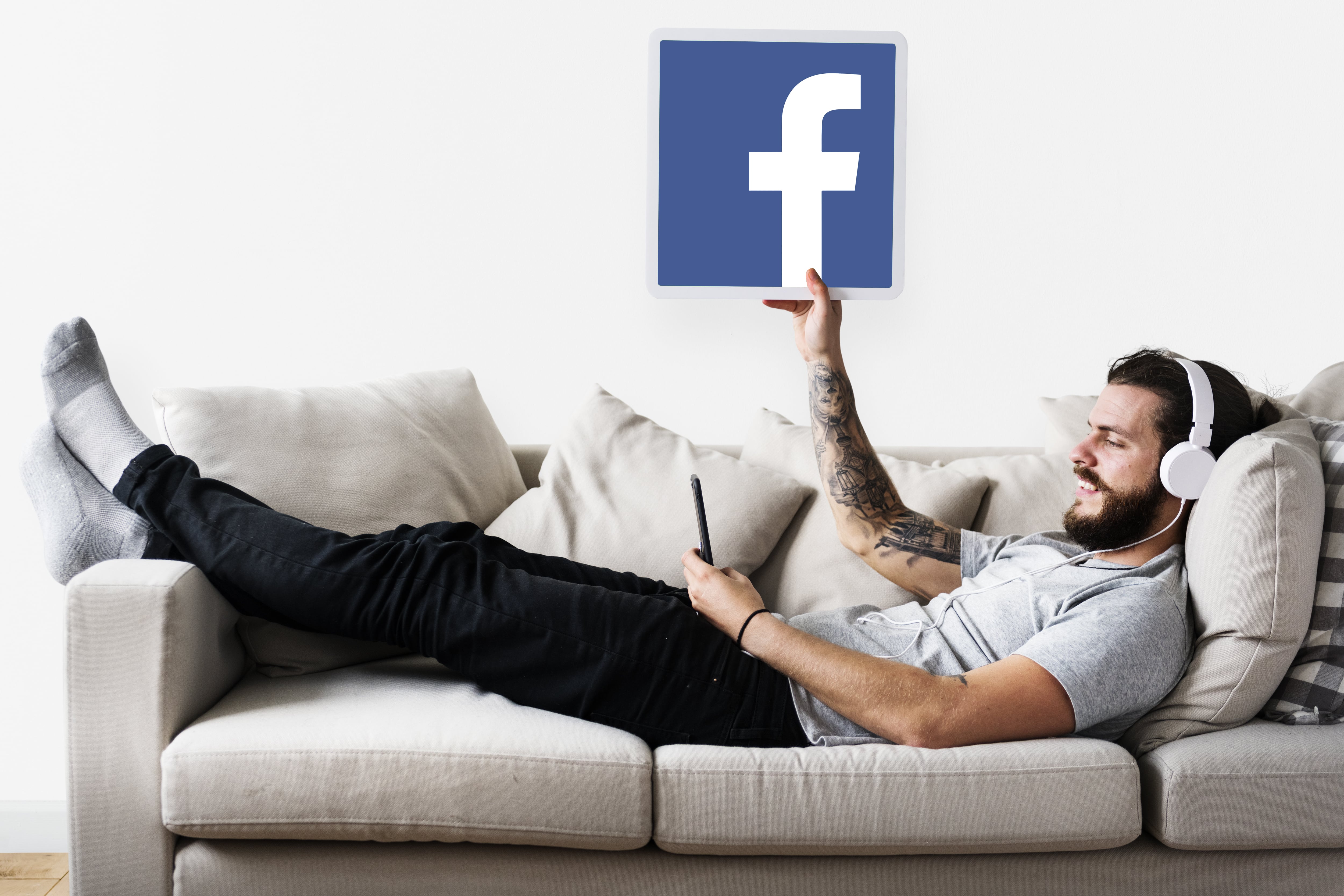 Mann auf Sofa mit Handy und Kopfhörern, hält Facebook-Logo. Beitrag über Entspannung beim Social-Media-Surfen.